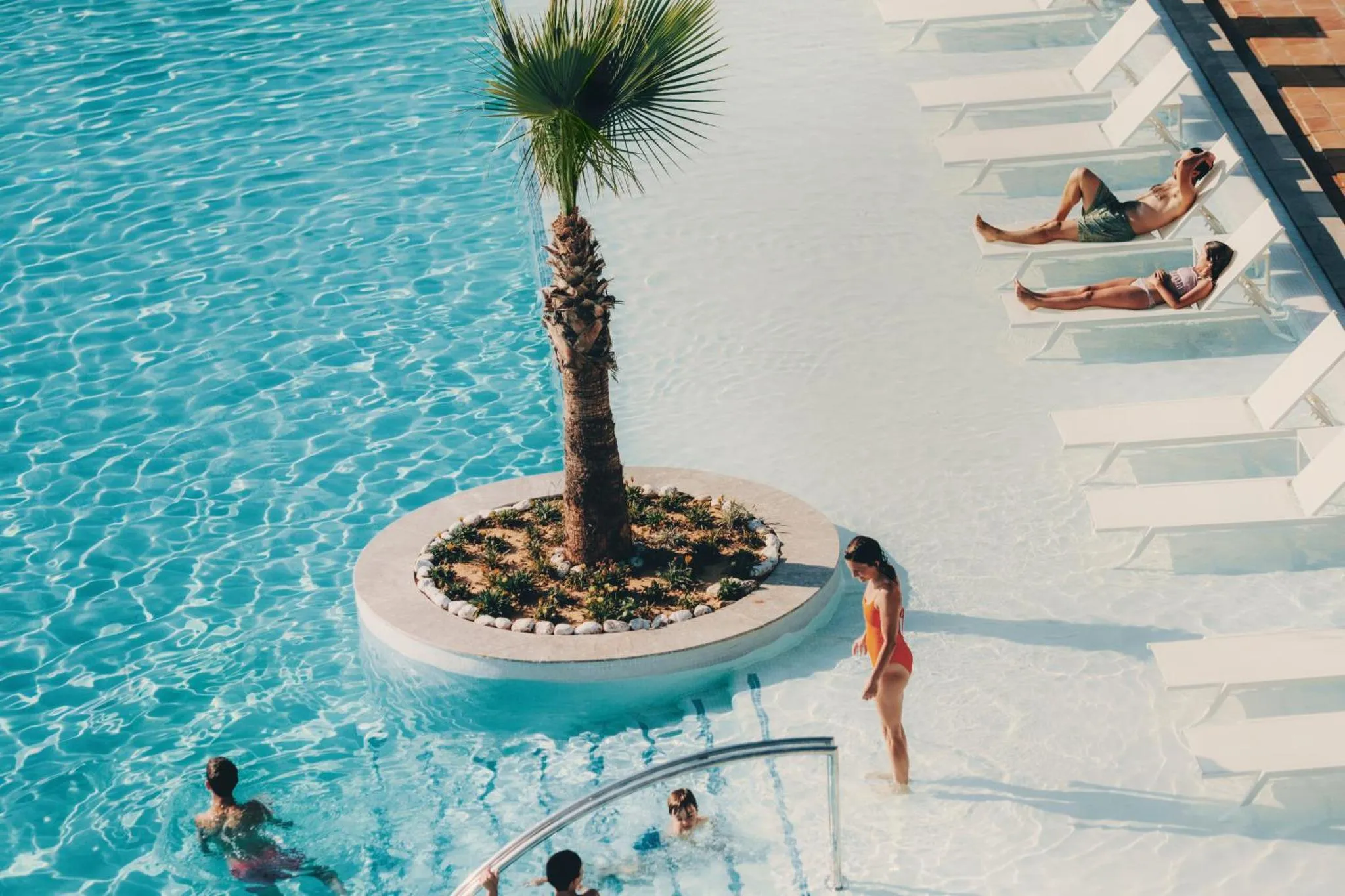 Des transats blancs bordent une piscine turquoise où se baignent quelques personnes.