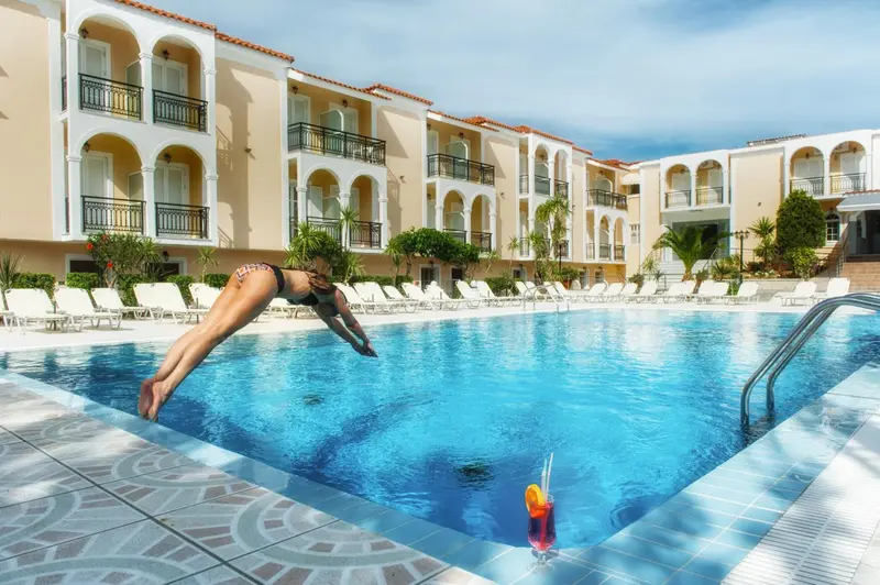 Une personne plonge dans une piscine ensoleillée devant un bâtiment beige.