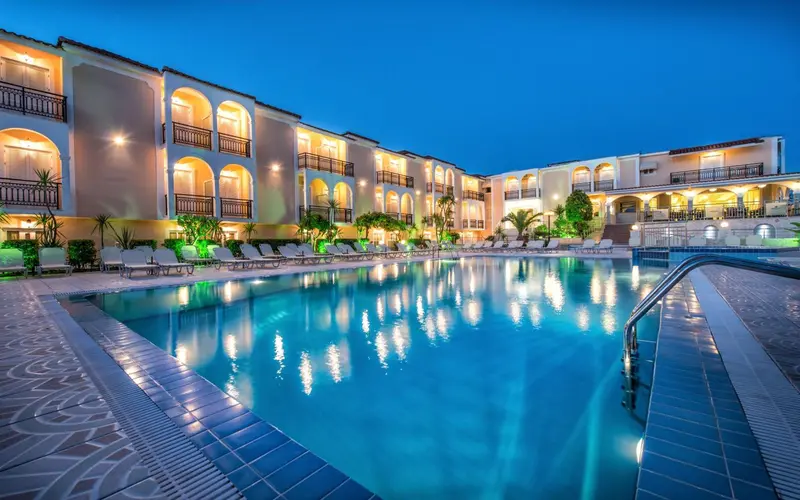 Piscine illuminée devant un bâtiment à plusieurs étages avec des balcons.