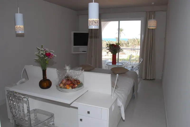 Une chambre lumineuse avec un lit et une table décorée de fleurs, donnant sur une vue mer.