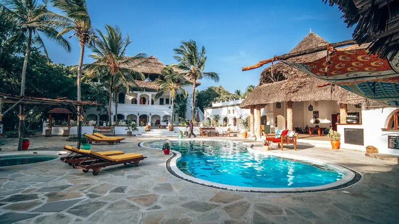Une piscine turquoise au centre d'une cour pavée, entourée de bâtiments blancs et de végétation tropicale.