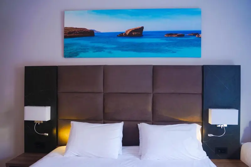 Un lit double avec une tête de lit matelassée face à une grande toile représentant une côte rocheuse et la mer.