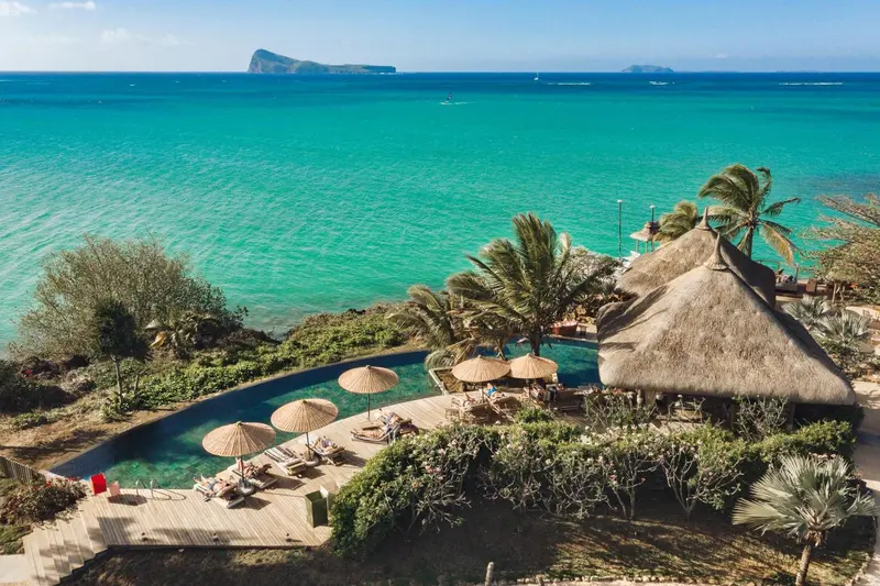 Vue aérienne d'une piscine bordée de transats et de palmiers, avec la mer turquoise en arrière-plan.