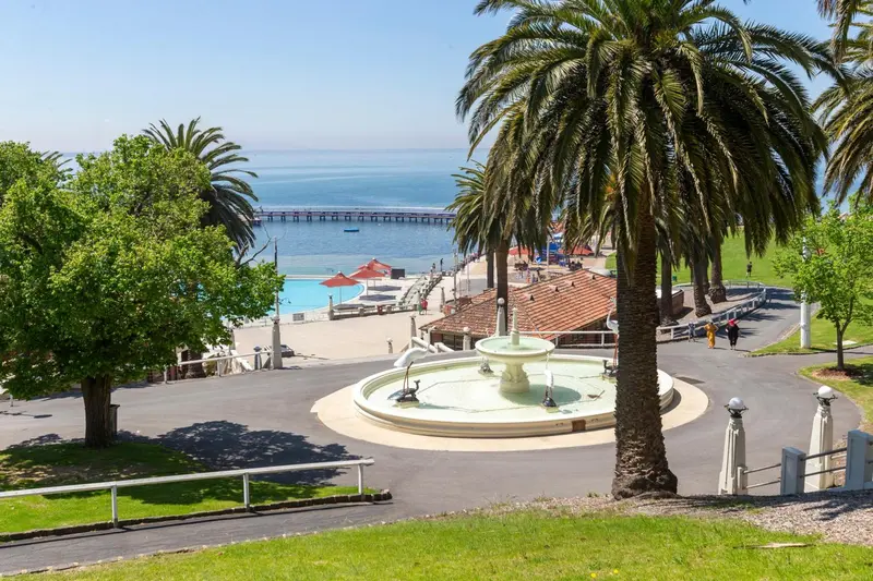 Vue d'un jardin avec une fontaine et des palmiers surplombant une plage et une jetée.