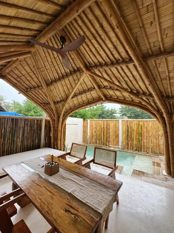 Une table en bois est placée sous une structure en bambou ouverte sur une piscine.