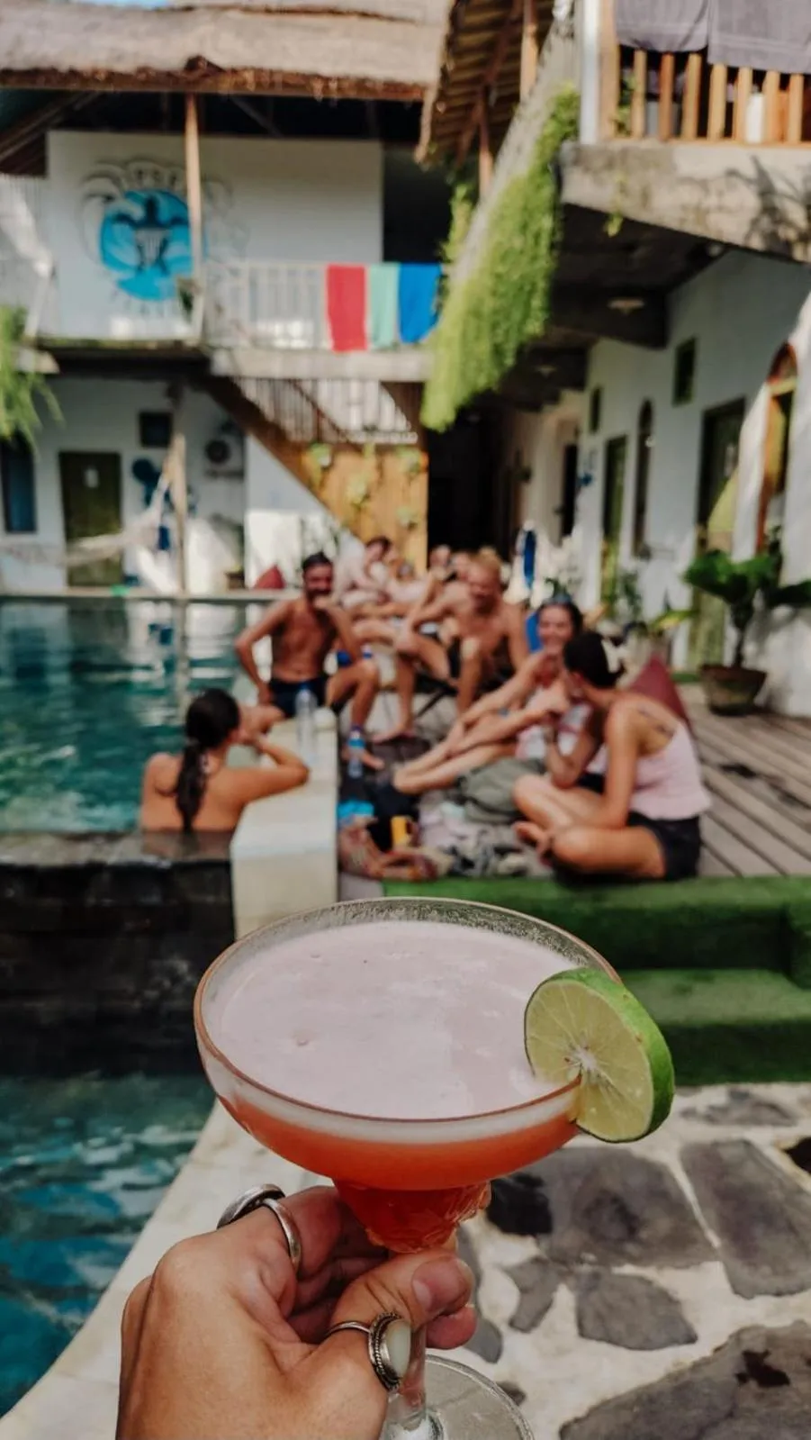 Un cocktail coloré est tenu au premier plan, avec une piscine et des personnes assises en arrière-plan.