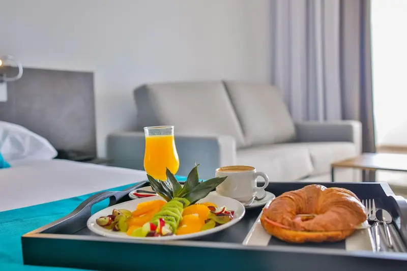 Un plateau de petit-déjeuner avec des fruits frais, une viennoiserie et des boissons est présenté sur un lit.