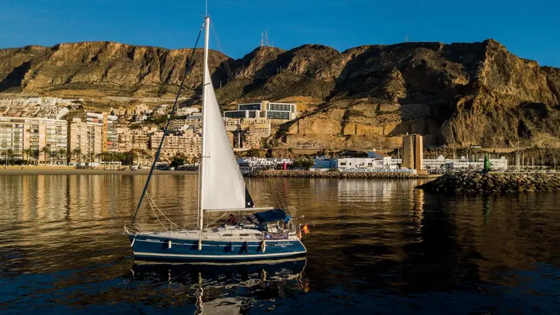 Un voilier navigue sur une eau calme avec une côte montagneuse en arrière-plan.