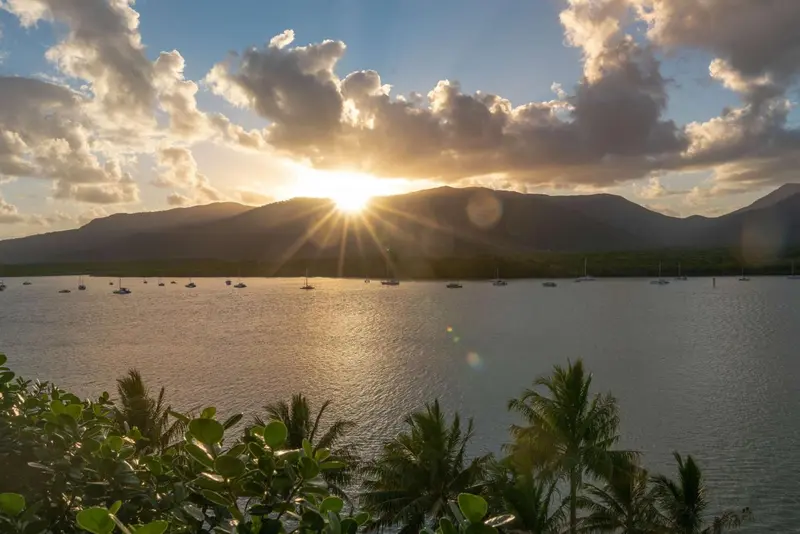 Coucher de soleil sur une baie avec des bateaux amarrés et une végétation tropicale au premier plan.