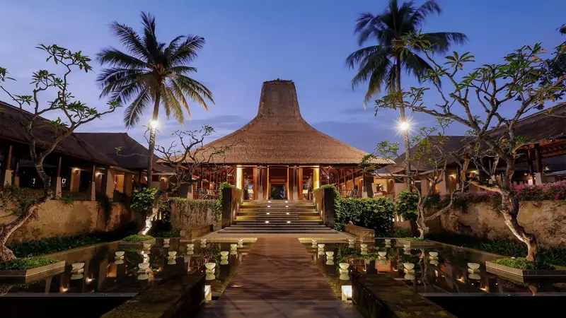 Bâtiment à toit de chaume éclairé au crépuscule, avec des bassins d'eau et une végétation tropicale.