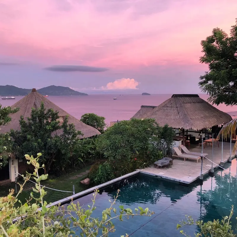 Deux toits de chaume surplombent une piscine et la mer au coucher du soleil.