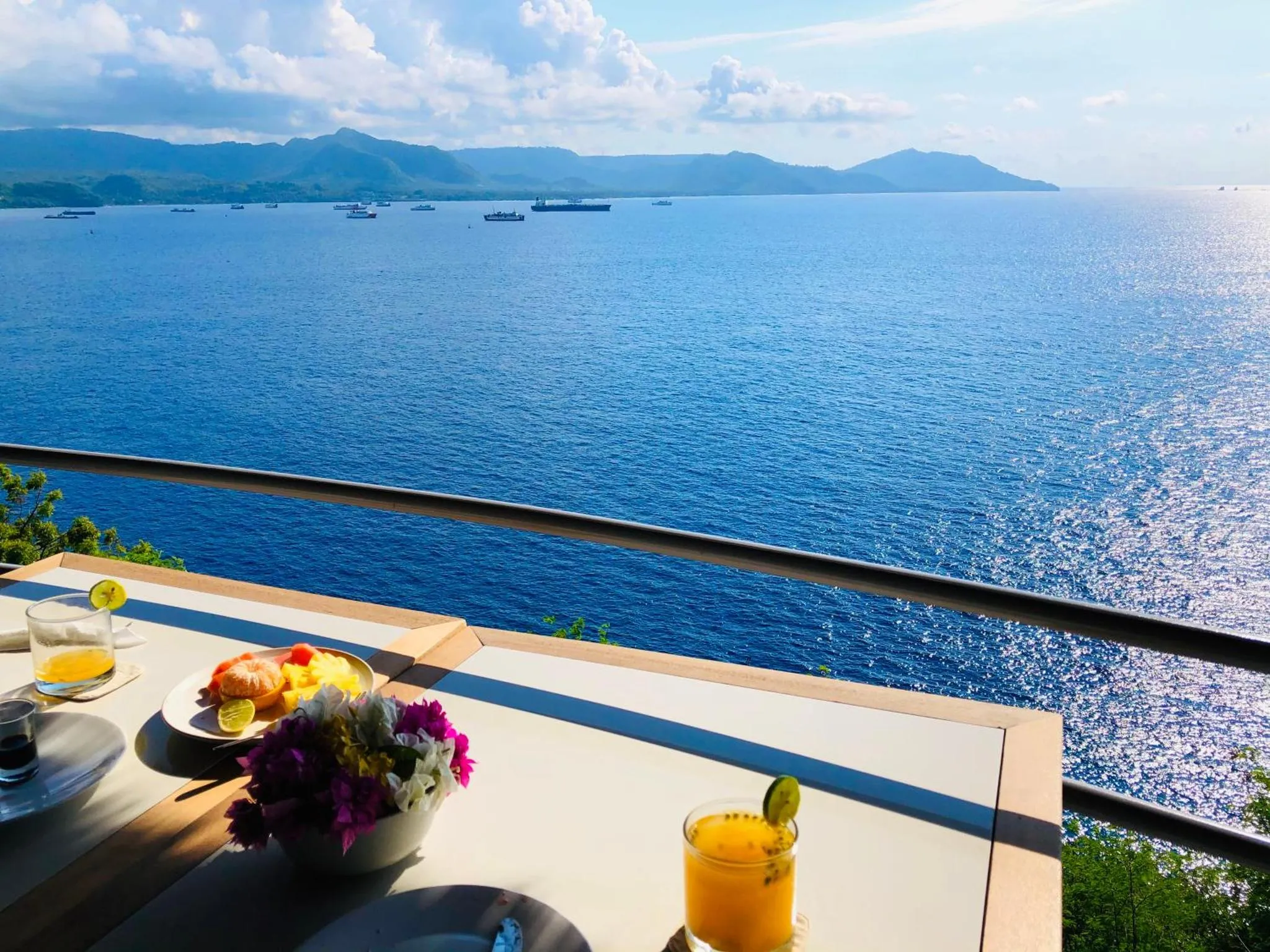 Une table est dressée avec des boissons et des fruits sur une terrasse surplombant la mer.