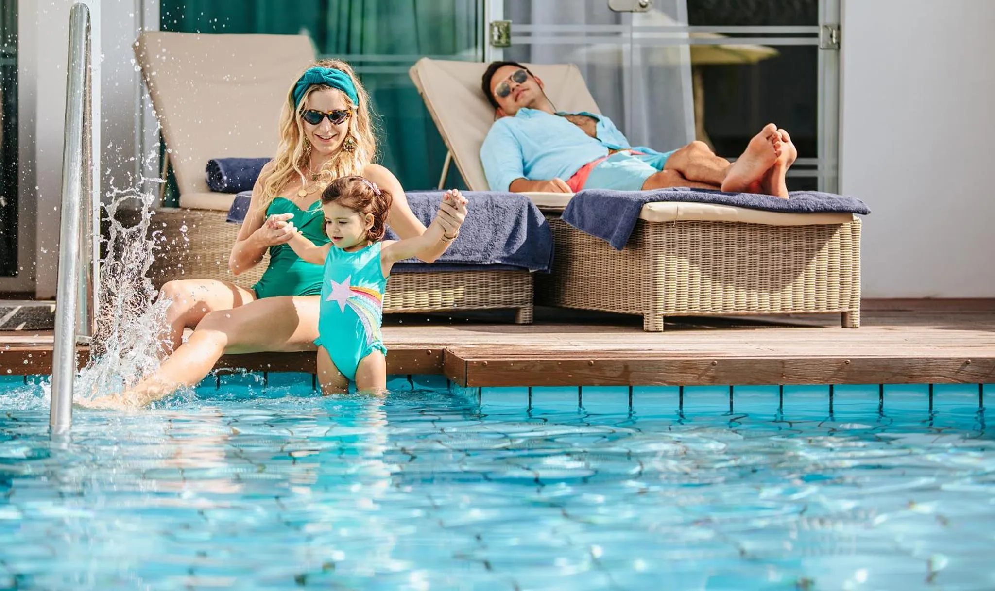 Une femme et un enfant s'amusent dans une piscine avec un homme relaxant sur des transats.