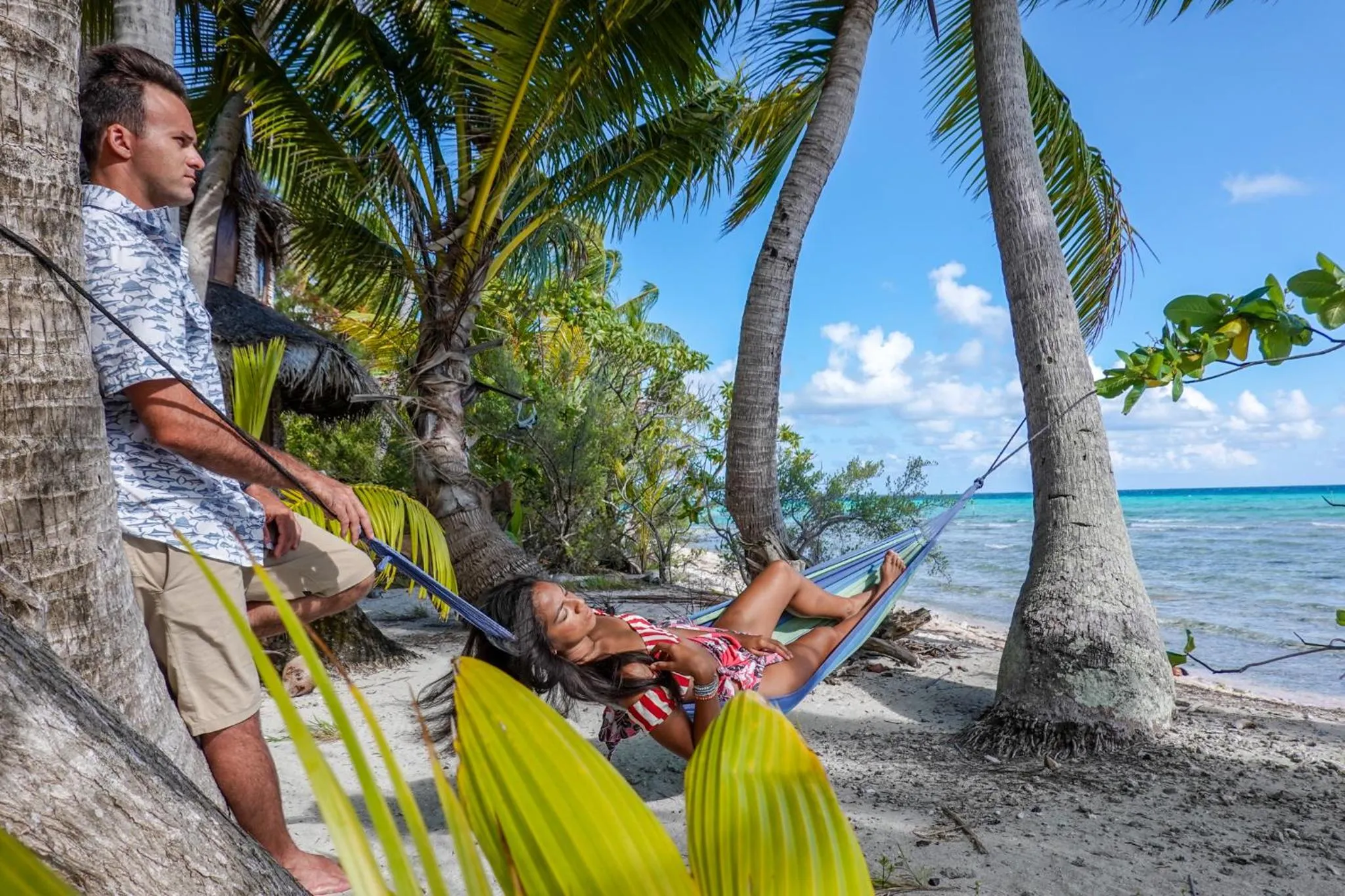 Deux personnes se détendent dans des hamacs sur une plage de sable blanc bordée de palmiers.