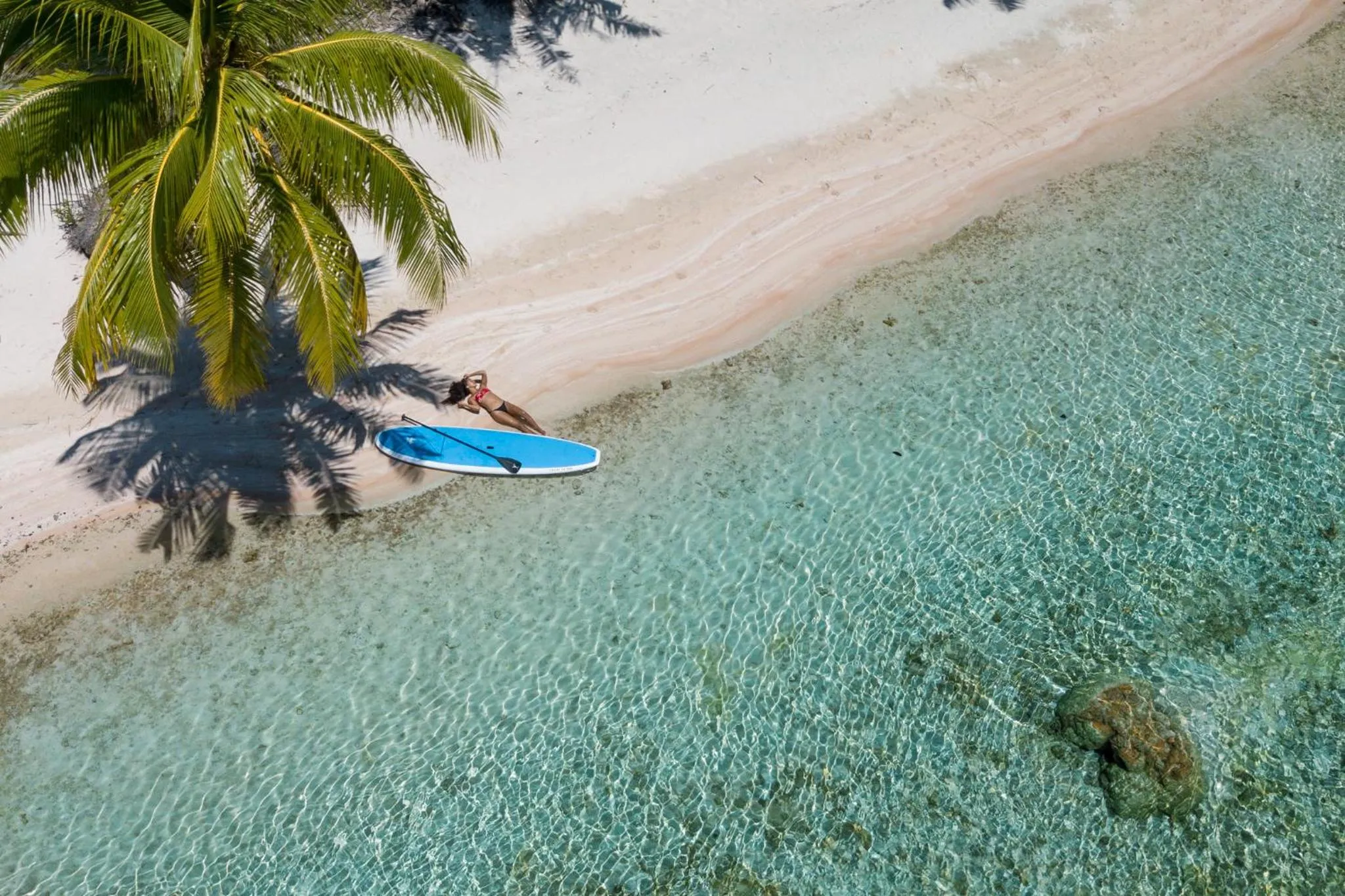 Une personne se détend sur une planche de paddle dans une eau turquoise peu profonde le long d'une plage de sable blanc.