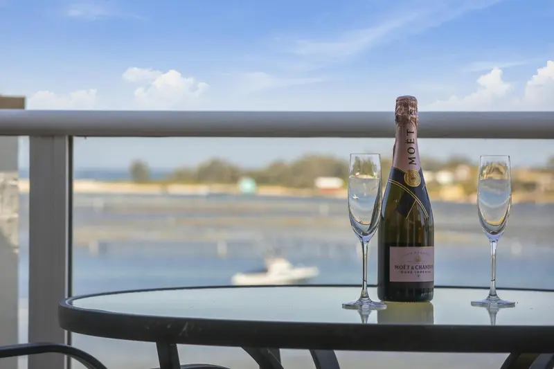 Une table ronde en verre avec une bouteille de champagne et deux coupes, sur une terrasse avec vue sur la mer.