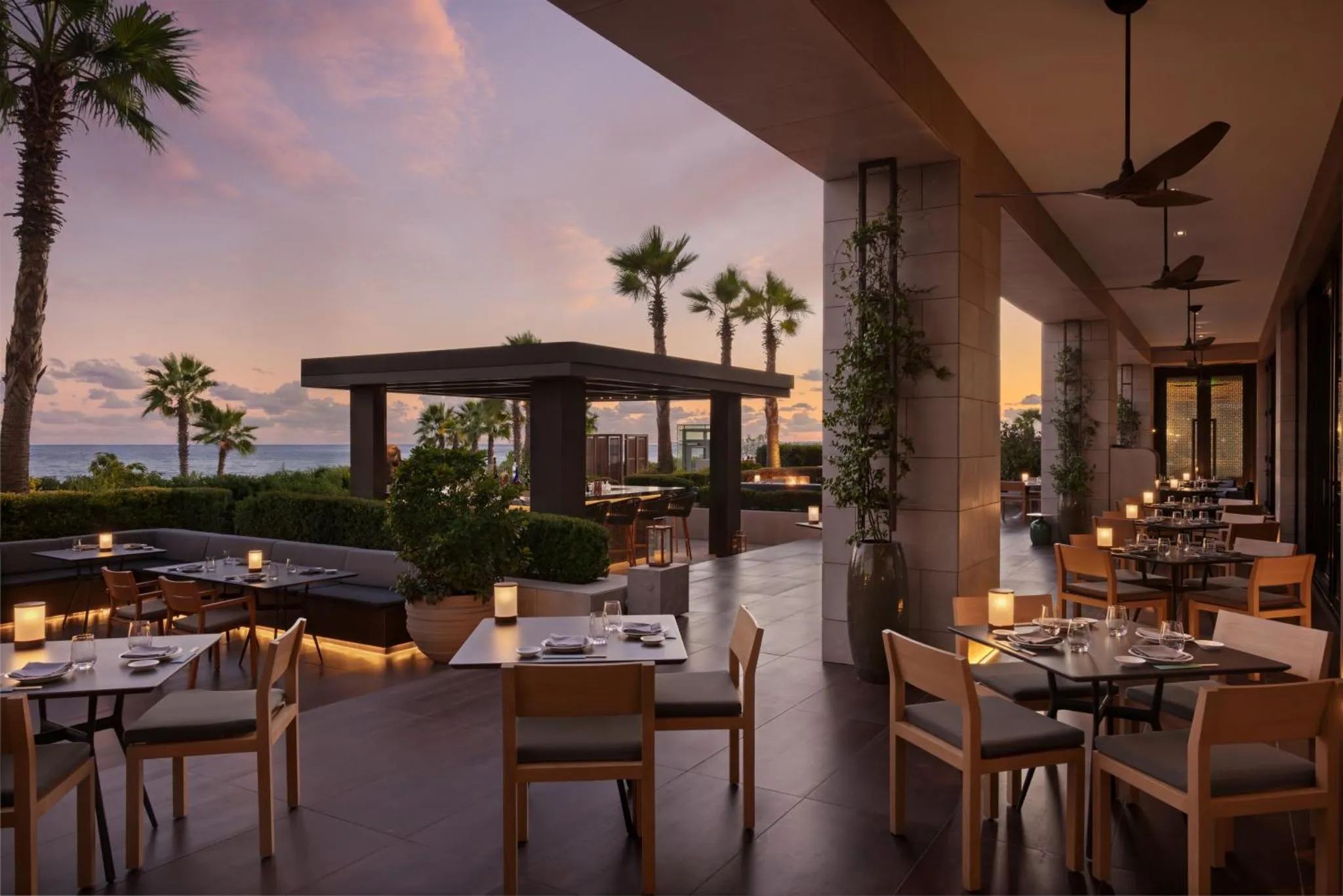 Terrasse de restaurant avec tables dressées face à la mer au coucher du soleil.