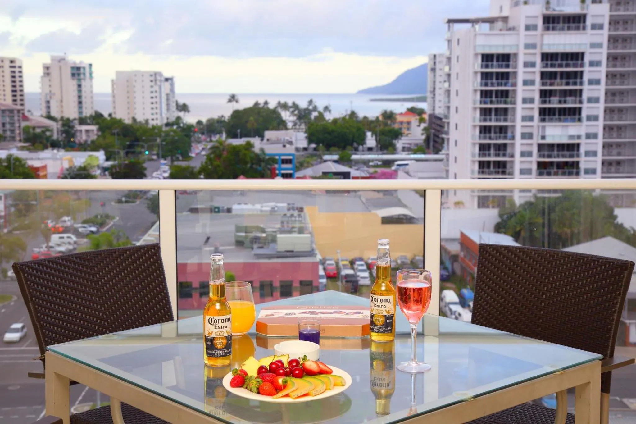 Une table basse en verre avec des boissons et des fruits est mise en scène sur une terrasse.