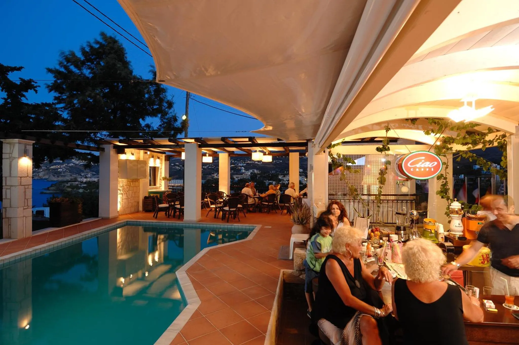 Une terrasse illuminée surplombe une piscine et offre une vue sur la mer au loin.