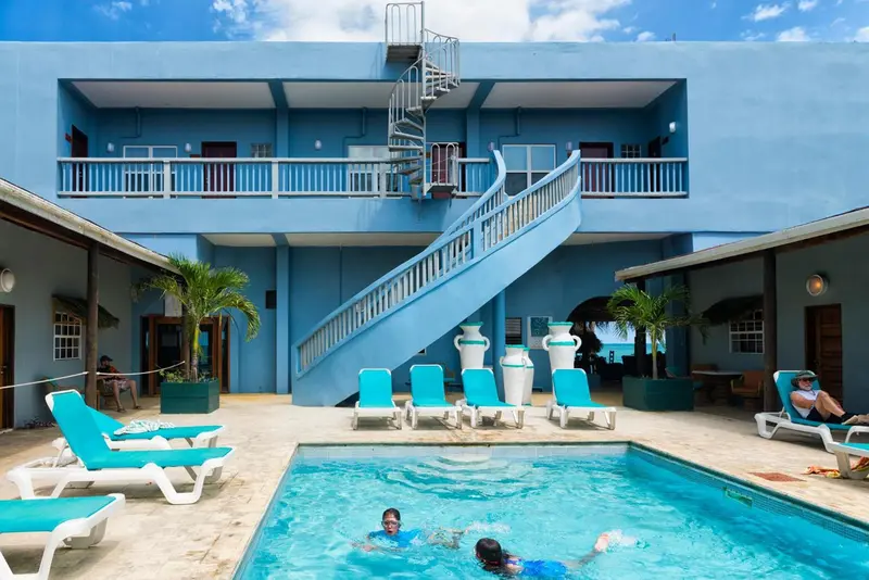 Une piscine turquoise est entourée de chaises longues bleues et de palmiers, devant un bâtiment bleu clair avec un escalier en colimaçon.