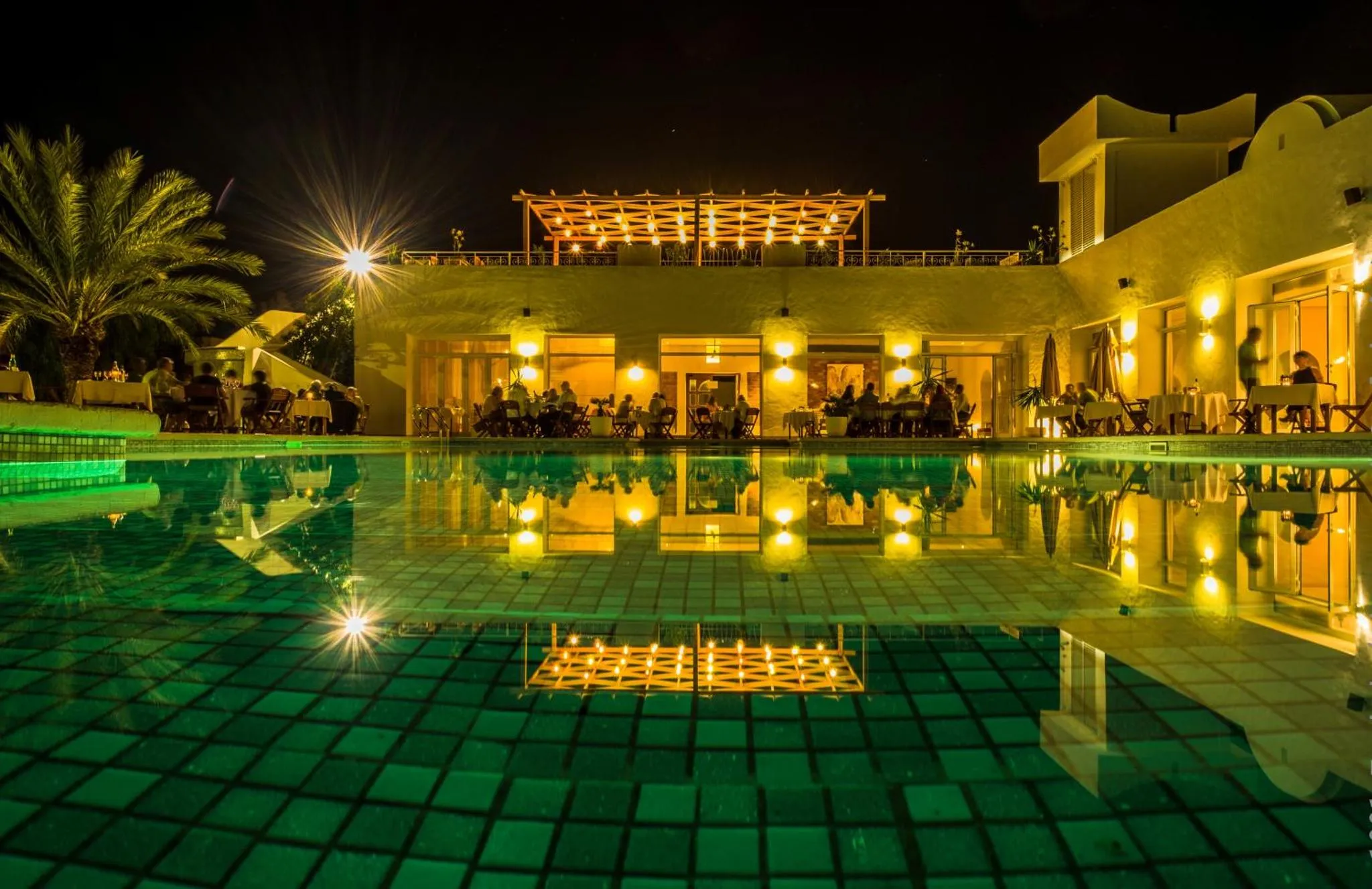 Une piscine illuminée reflète les lumières d'un bâtiment et d'une terrasse.