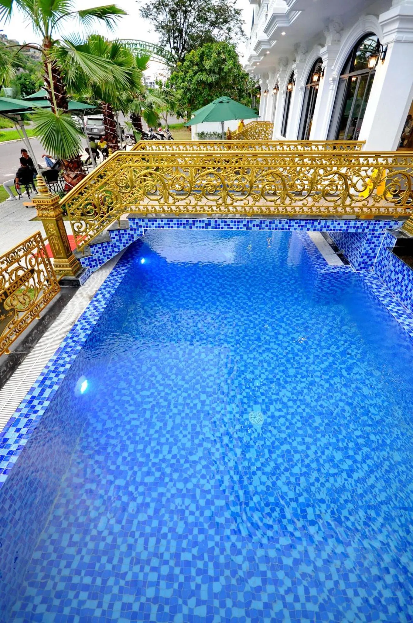 Une piscine rectangulaire aux carreaux bleus est entourée d'une terrasse et d'une balustrade dorée.