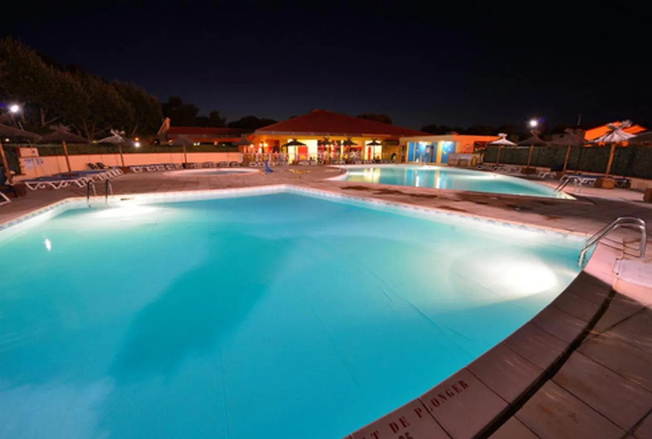 Piscine illuminée au crépuscule avec transats et bâtiments en arrière-plan.