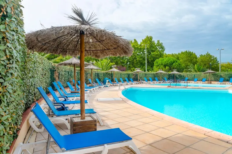 Transats bleus alignés au bord d'une piscine ensoleillée sous des parasols en chaume.