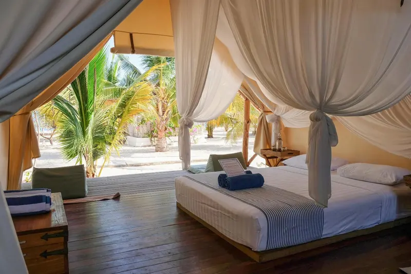 Un lit recouvert de moustiquaire dans une chambre ouverte sur une plage de sable blanc.