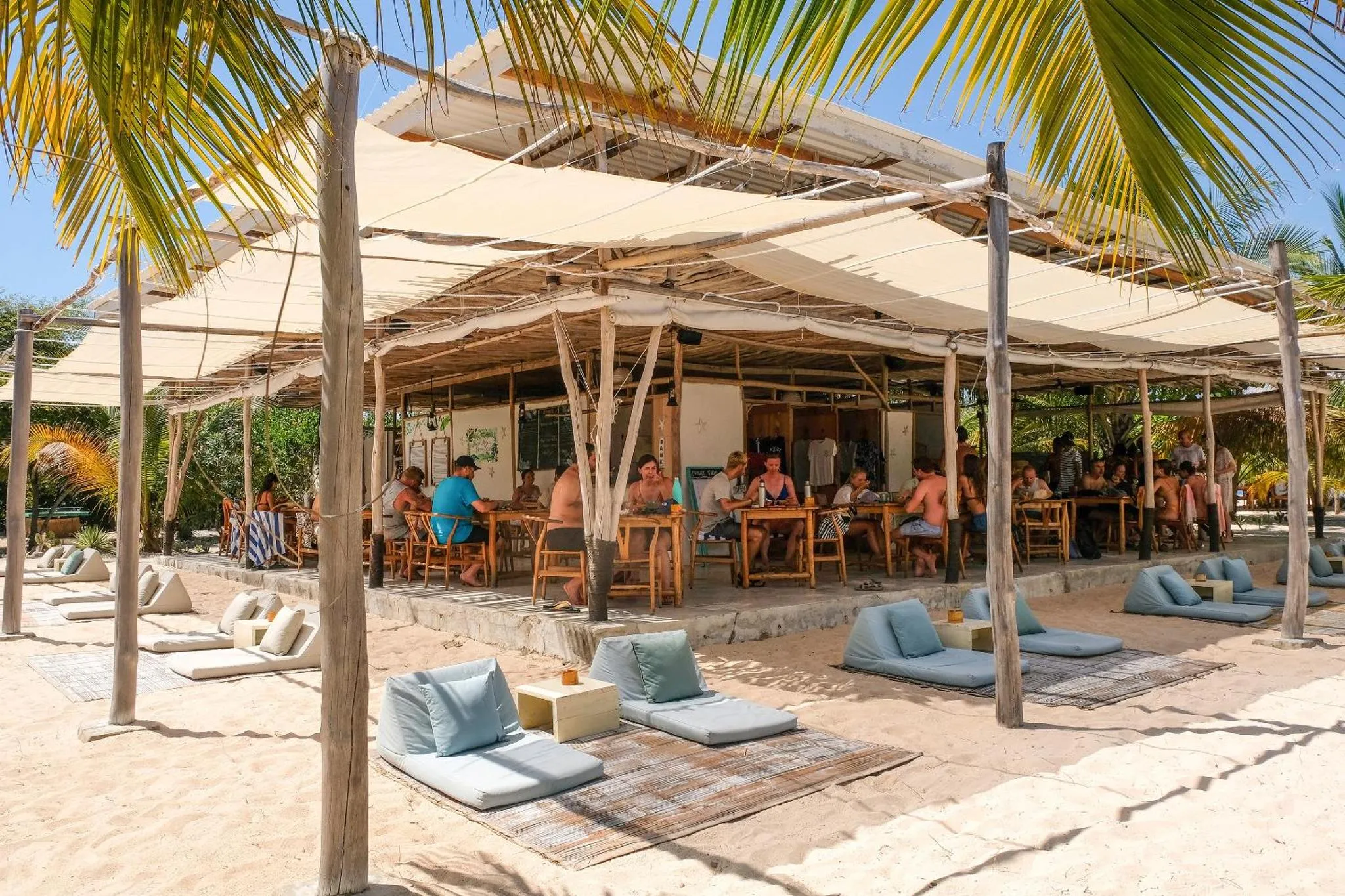 Terrasse ombragée avec tables et chaises, donnant sur une plage de sable.