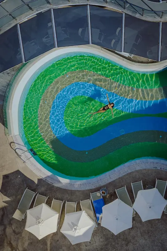 Vue aérienne d'une piscine circulaire aux eaux turquoise et verdoyantes, entourée de chaises longues blanches.