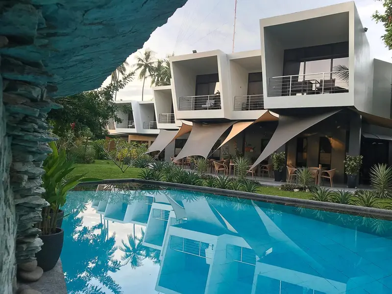 Bâtiment moderne blanc avec des balcons surplombant une piscine turquoise et une pelouse verdoyante.