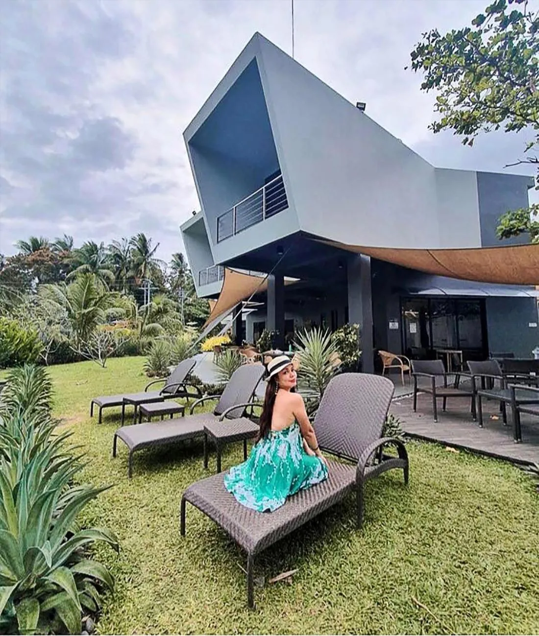 Une personne assise sur un transat dans un jardin verdoyant devant un bâtiment moderne.