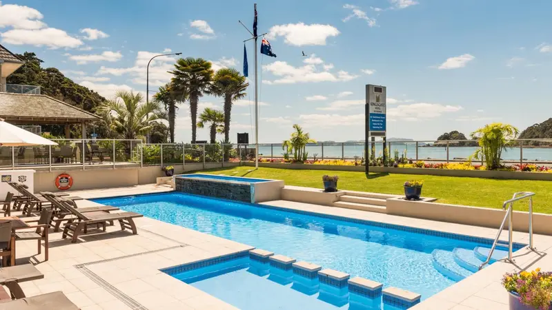 Une piscine rectangulaire avec des marches et des transats, devant un bâtiment blanc et une pelouse verdoyante.