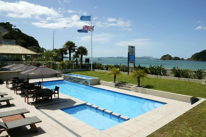 Une piscine rectangulaire avec des transats et des parasols, devant un bâtiment et une mer bleue.