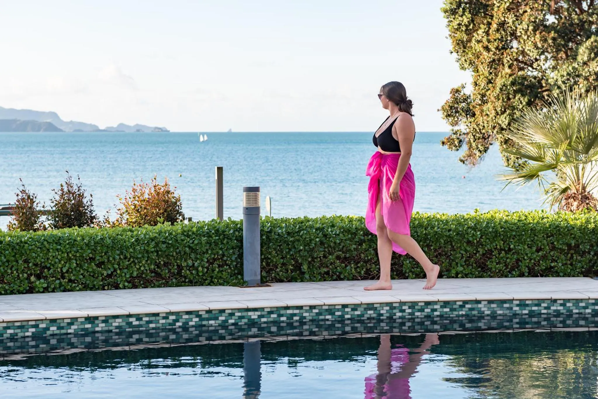 Une femme traverse un espace pavé près d'une piscine avec une vue sur la mer.