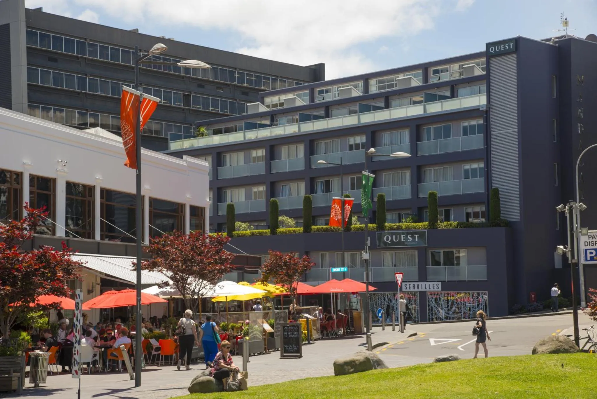 Terrasse ensoleillée avec parasols rouges et tables de restaurant devant un bâtiment moderne.