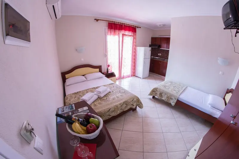 Une chambre lumineuse avec un lit double et un lit simple, une kitchenette et une table avec des fruits.