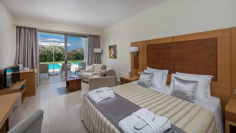Chambre lumineuse avec un grand lit et un coin salon donnant sur une piscine.
