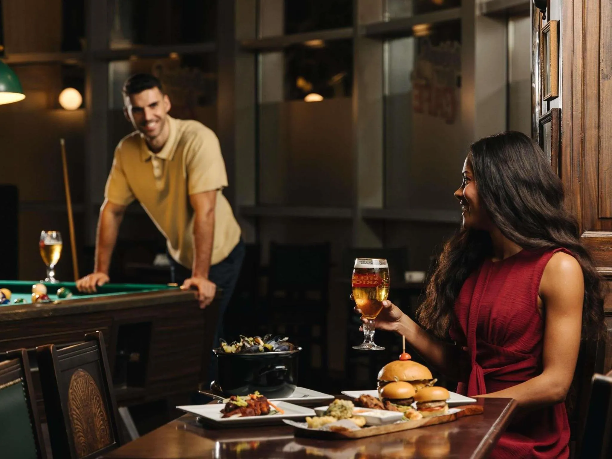 Deux clients savourent des boissons et des amuse-gueules dans un bar avec un billard en arrière-plan.