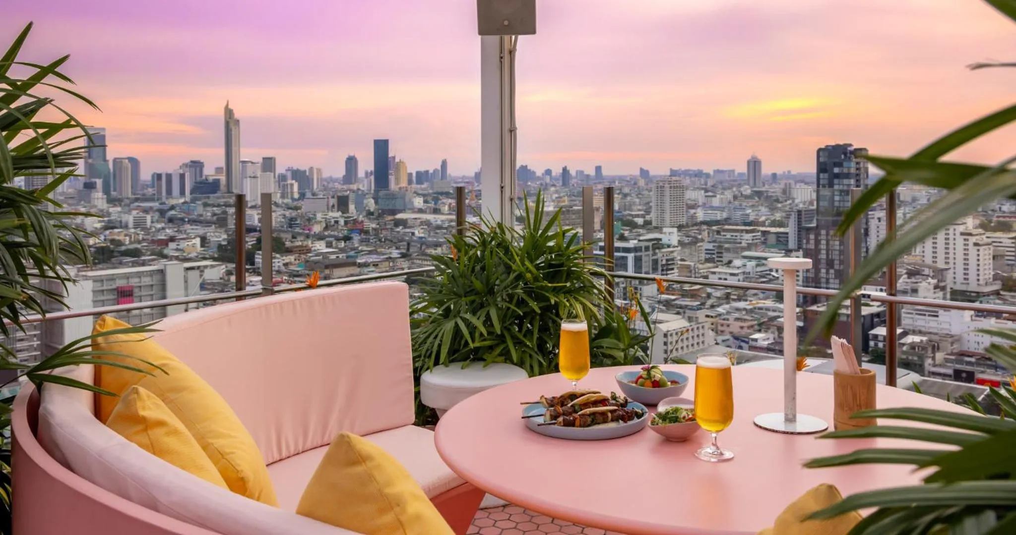 Une table rose avec des boissons et des plats est mise en scène sur une terrasse avec vue sur la ville au coucher du soleil.