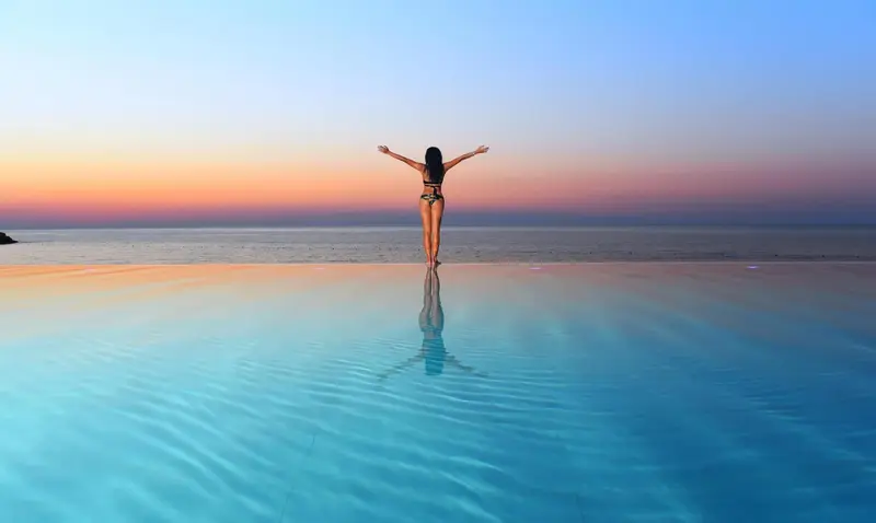 Une personne se tient debout, les bras ouverts, au bord d'une piscine à débordement face à la mer.