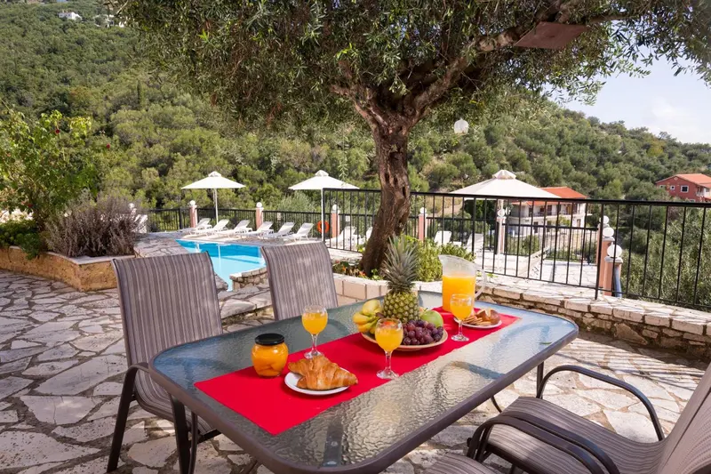 Une table de petit-déjeuner est dressée sur une terrasse ensoleillée avec vue sur une piscine et un paysage verdoyant.