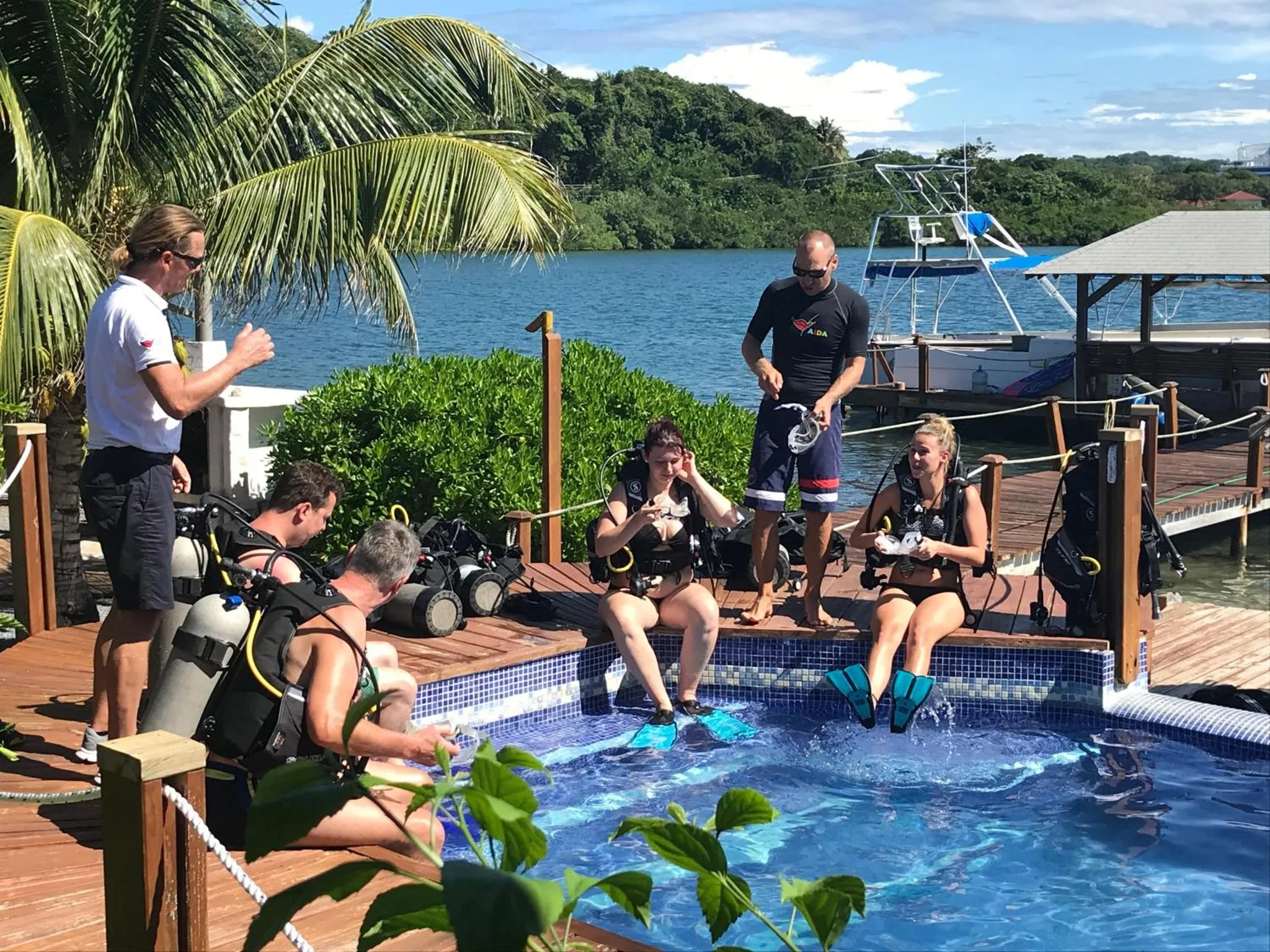 Des personnes équipées discutent au bord d'une piscine avec un ponton en arrière-plan.