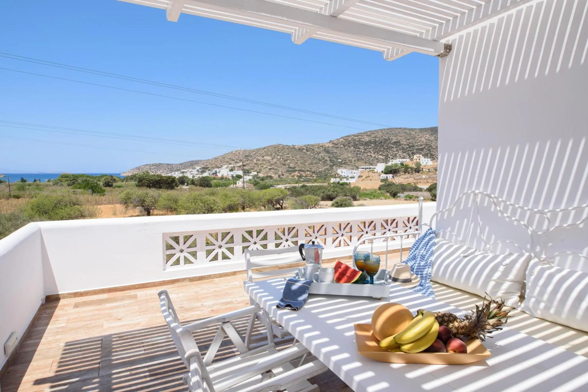 Une table et des chaises blanches sont disposées sur une terrasse ensoleillée avec vue sur la mer et les collines.