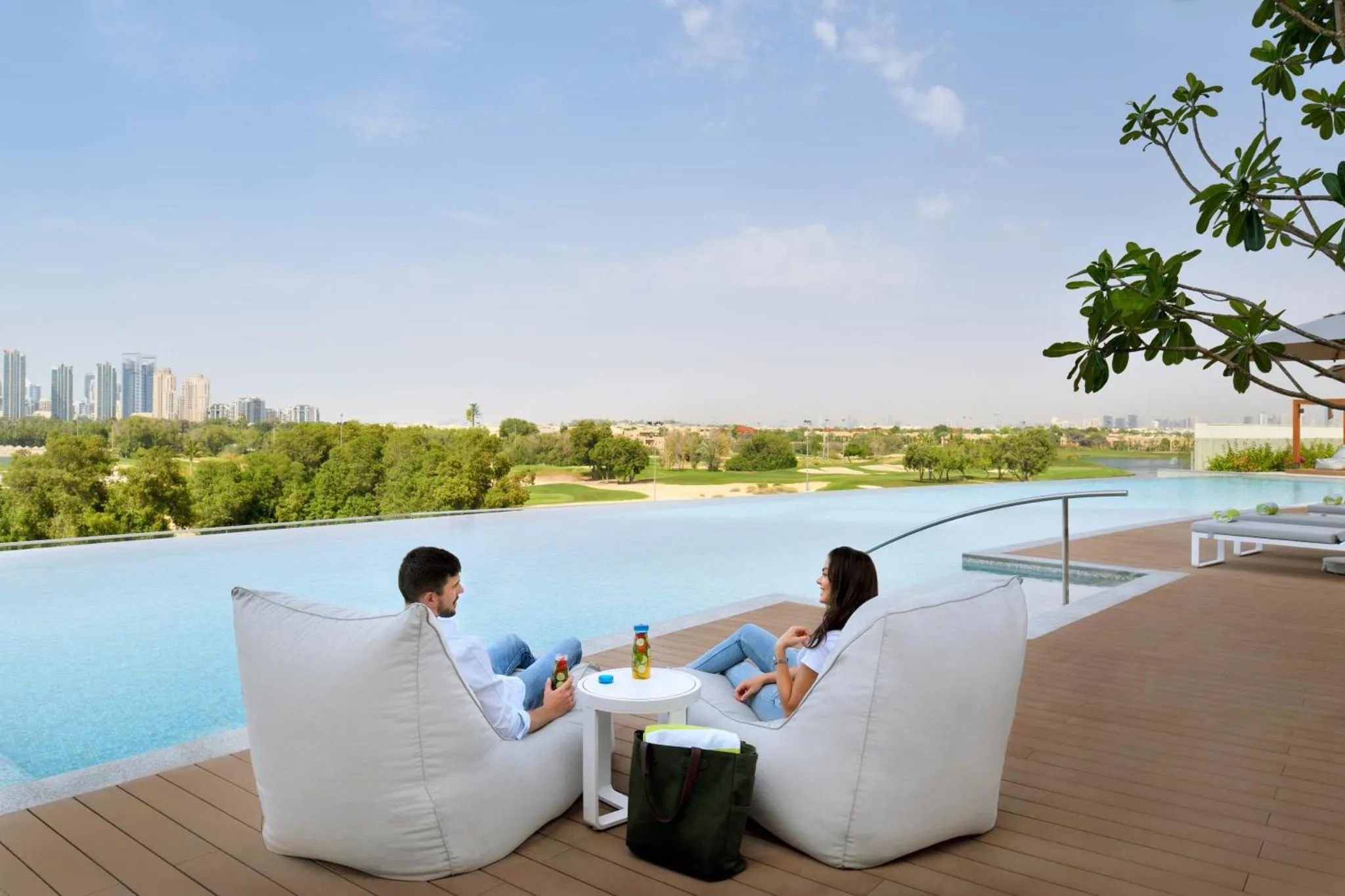 Deux personnes se détendent sur des fauteuils blancs au bord d'une piscine à débordement avec vue sur un paysage urbain.