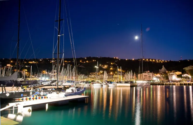 Port de plaisance illuminé au crépuscule avec des voiliers amarrés et des lumières scintillantes sur l'eau.