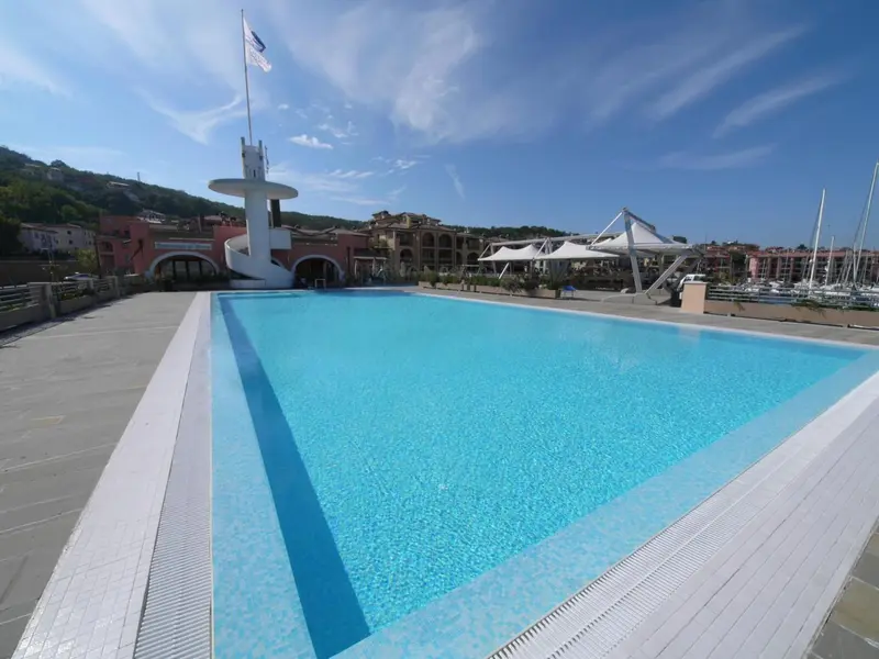 Une piscine rectangulaire turquoise au premier plan, avec un bâtiment et un port de plaisance en arrière-plan.