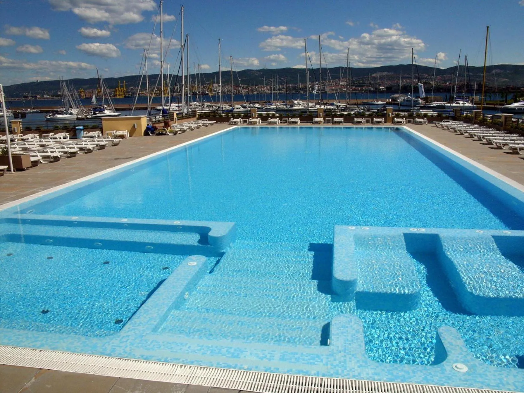 Une piscine rectangulaire bleue avec des marches et des transats blancs sur une terrasse ensoleillée.