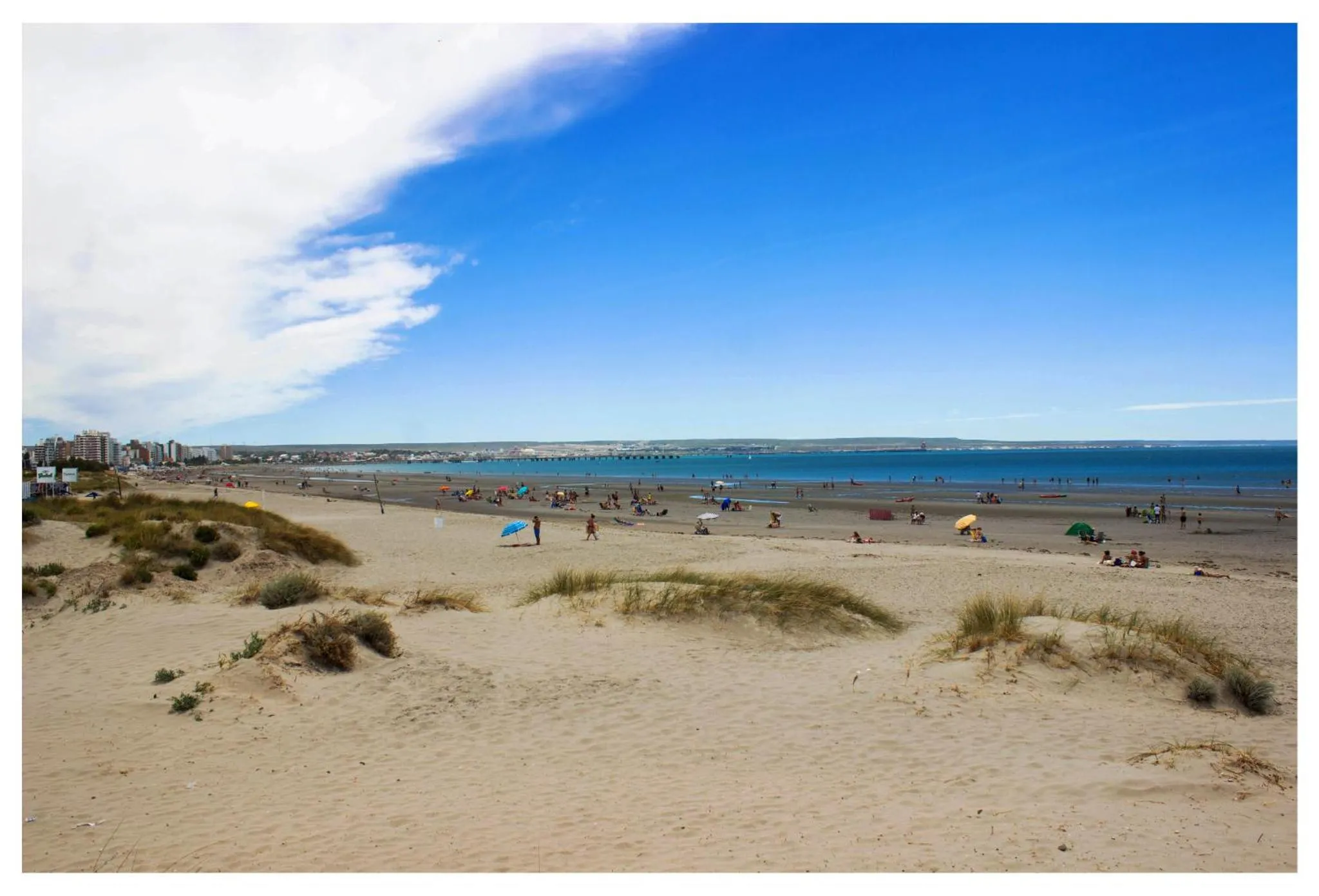 Plage de sable fin avec quelques personnes se baignant et des dunes en premier plan.